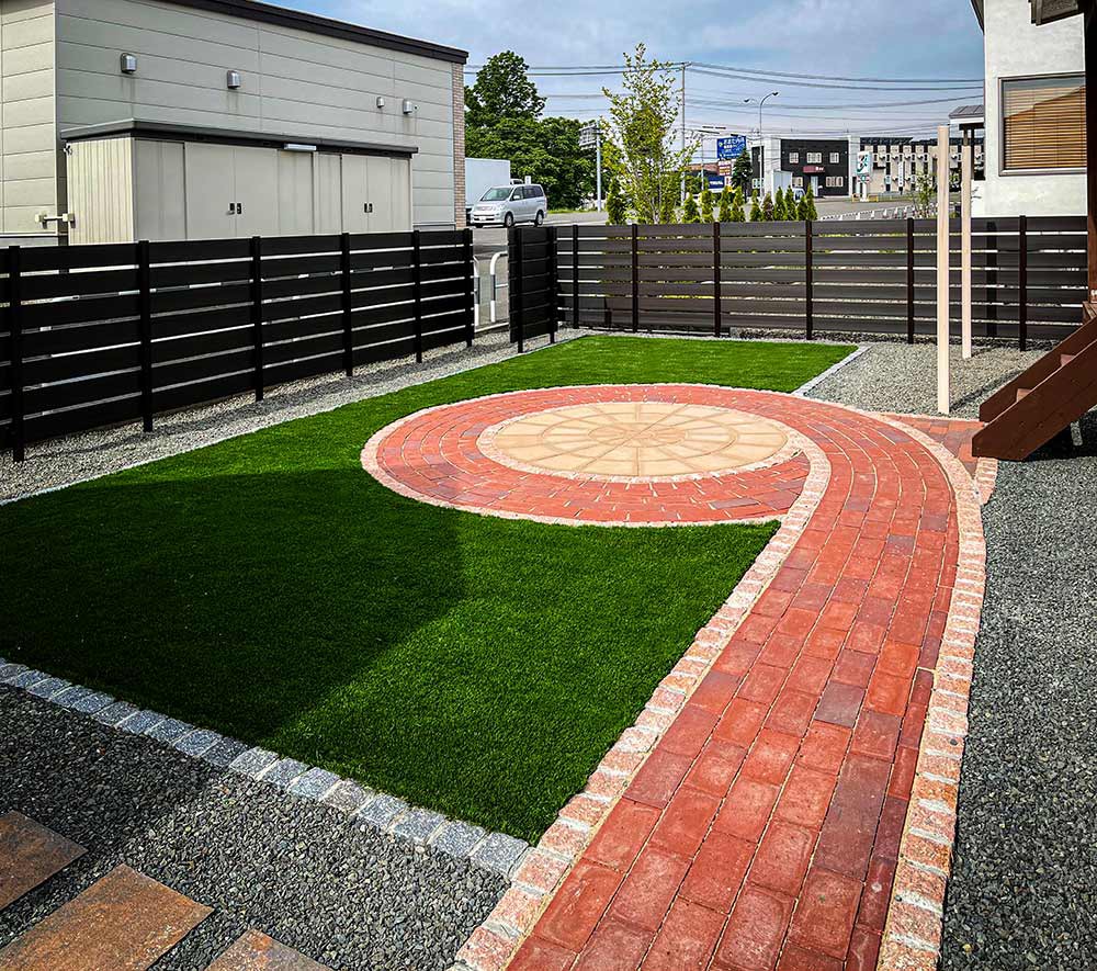 サークルペイバーをレンガで巻いた形状のガーデンテラスを中心に、人工芝生と人工木のフェンス、物干し台で構成された庭の景です。